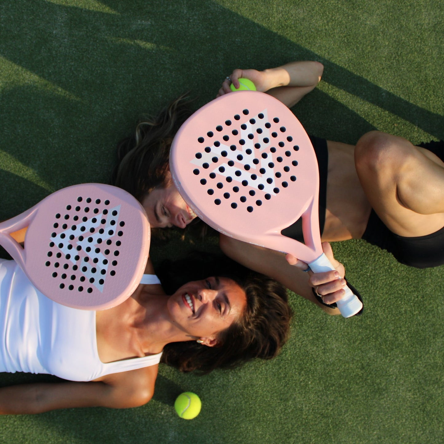 Two people lying on a tennis court with pink paddle rackets and yellow balls.
