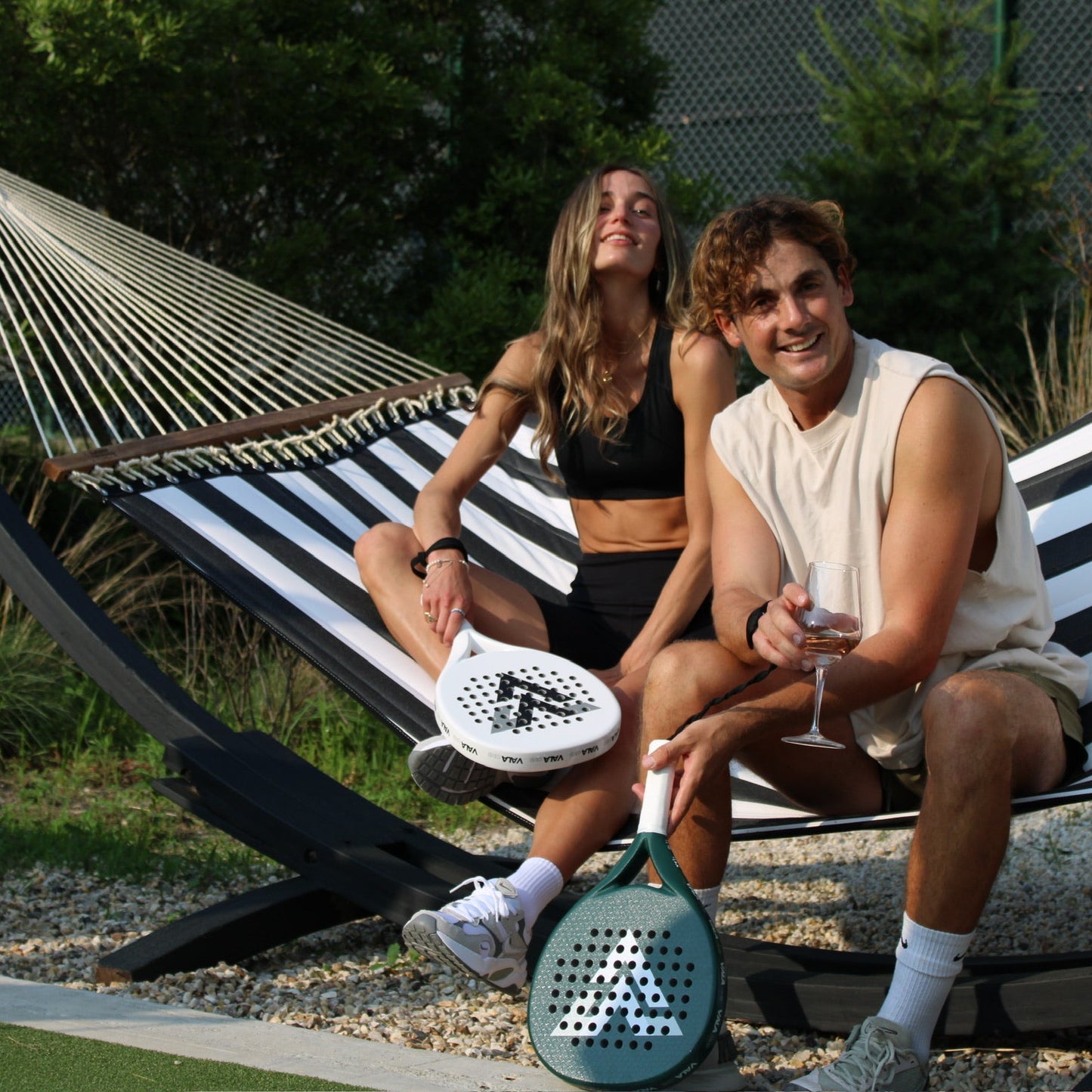 Two people sitting on a striped hammock with paddle rackets, surrounded by greenery.